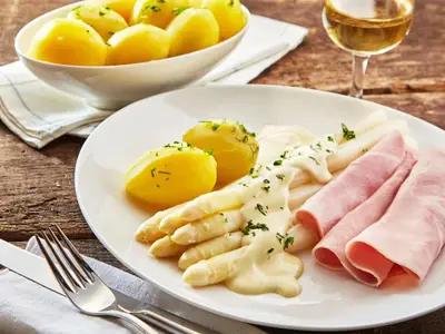 Delicious seasonal white asparagus with creamy mayonnaise, baby boiled potatoes and sliced ham or gammon served with white wine and utensils on a rustic table