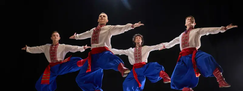 Four male dancers in formation, arms outstretched, dressed in embroidered shirts, blue trousers, and red boots, highlighting unity and tradition against black background. Concept of art and history.